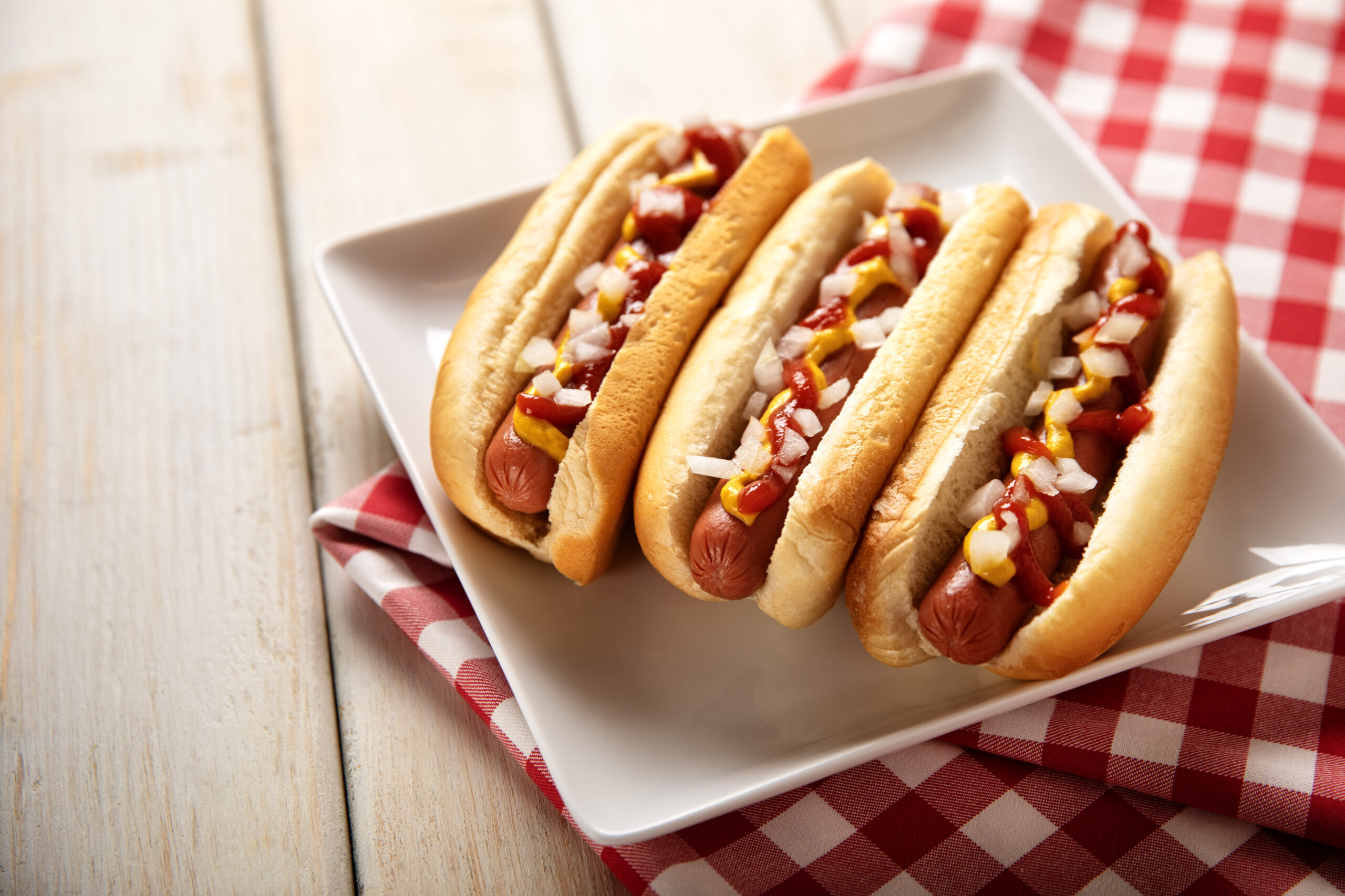 This is a photograph of three hot dogs on a white plate on a white wooden picnic bench. This is a great image for a Fourth of July picnic.