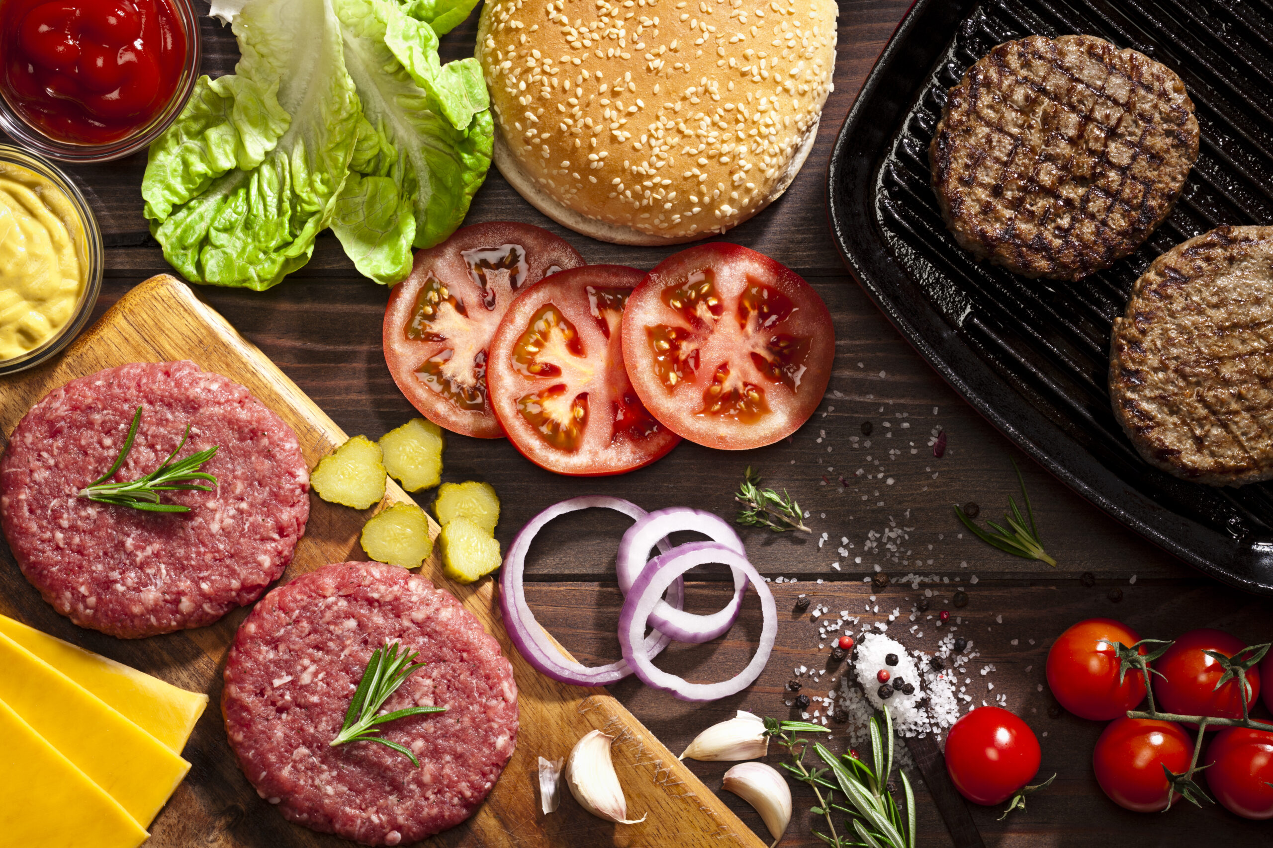 Top view of a rustic wood table filled with ingredients for cooking a delicious homemade cheeseburger. An iron grill with two cooked ground beef burgers is placed at the top-right of the frame and at the opposite side is a cutting board with raw ground beef burgers while the other ingredients are scattered on the table. DSRL studio photo taken with Canon EOS 5D Mk II and Canon EF 100mm f/2.8L Macro IS USM