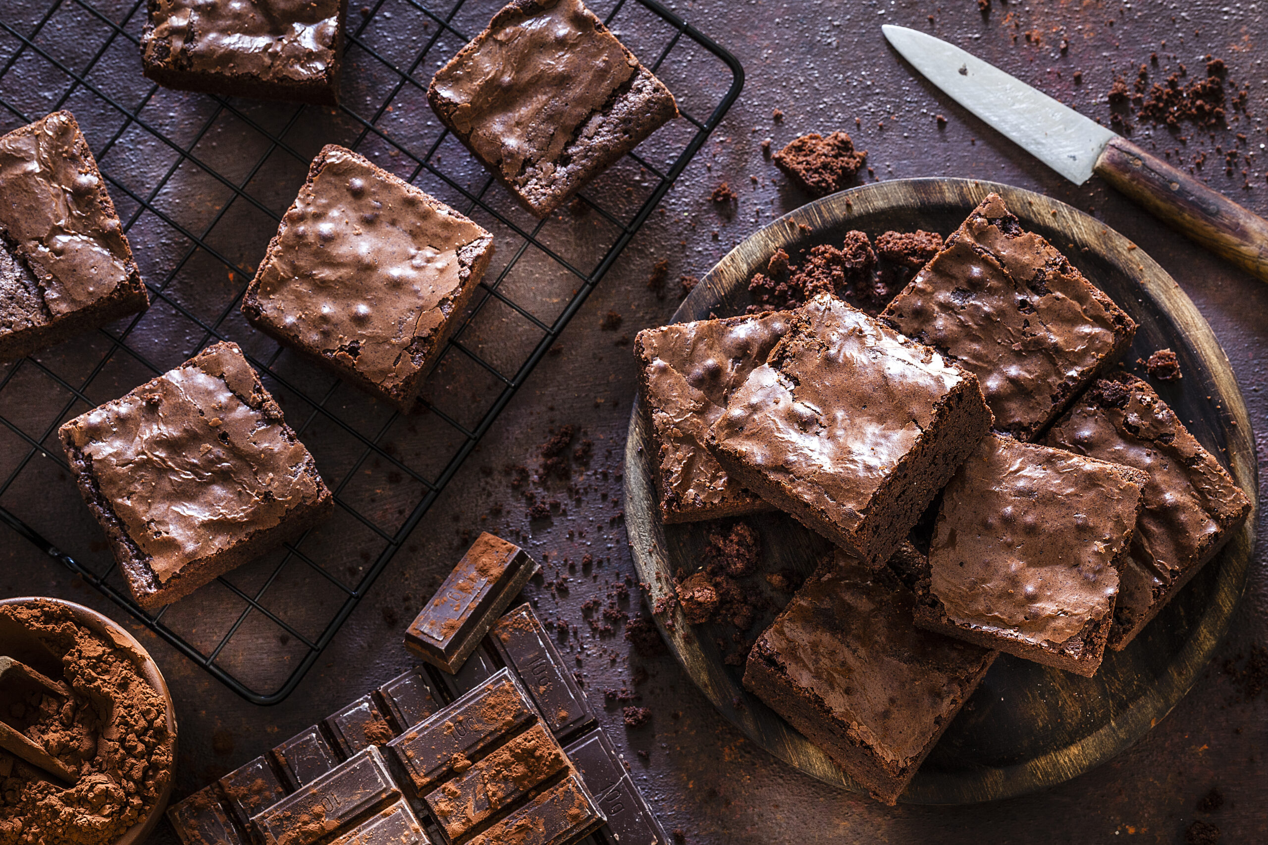 Top view of homemade chocolate brownies shot on dark table. A wooden plate filled with chocolate brownies is at the right and a cooling rack with brownies is at the left. Some chocolate bars are at the bottom. A brown bowl with cocoa powder is at the bottom left corner of an horizontal frame.