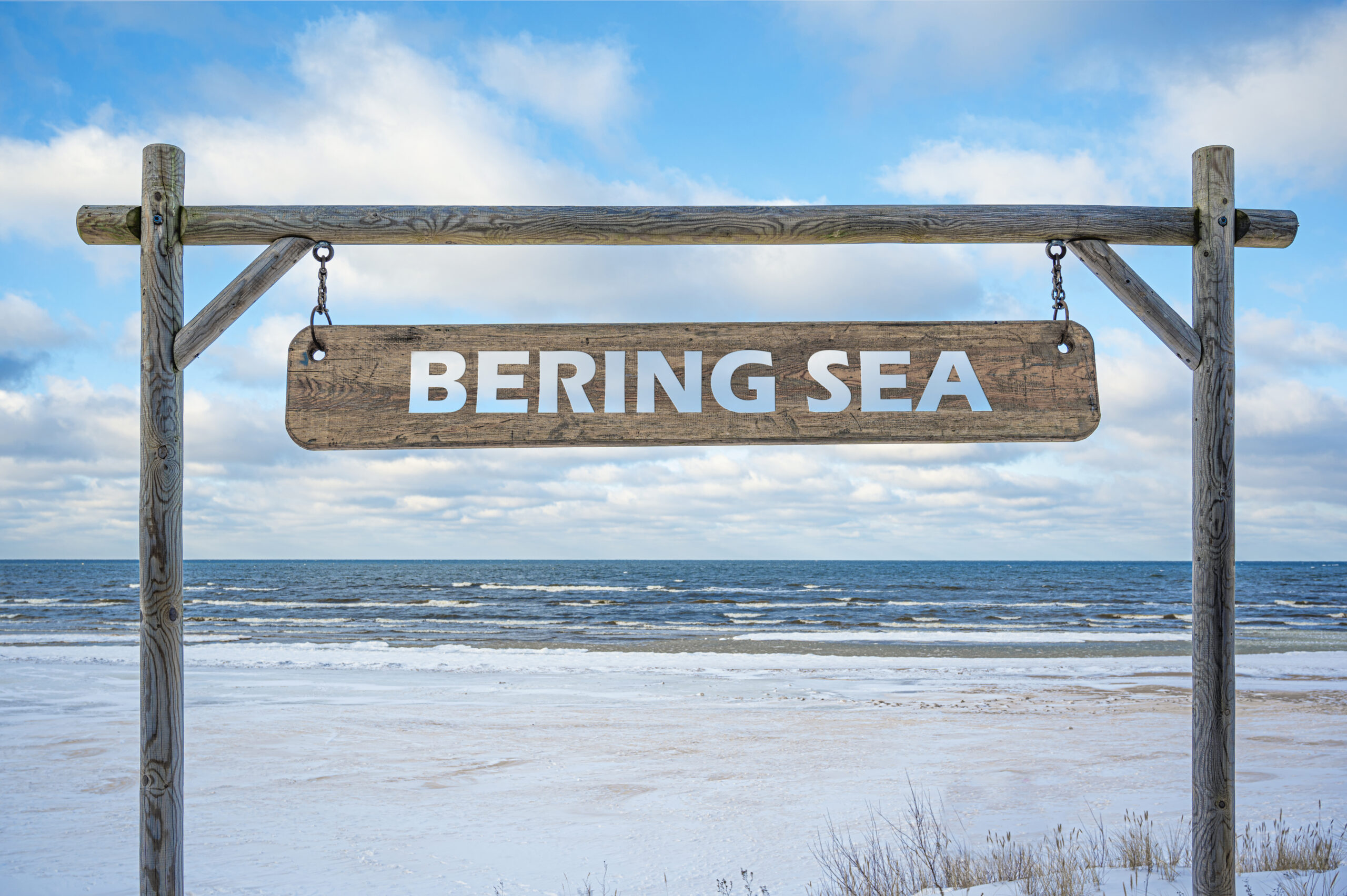 Wooden sign with Bering sea text against sea, blue sky and beach with white snow.