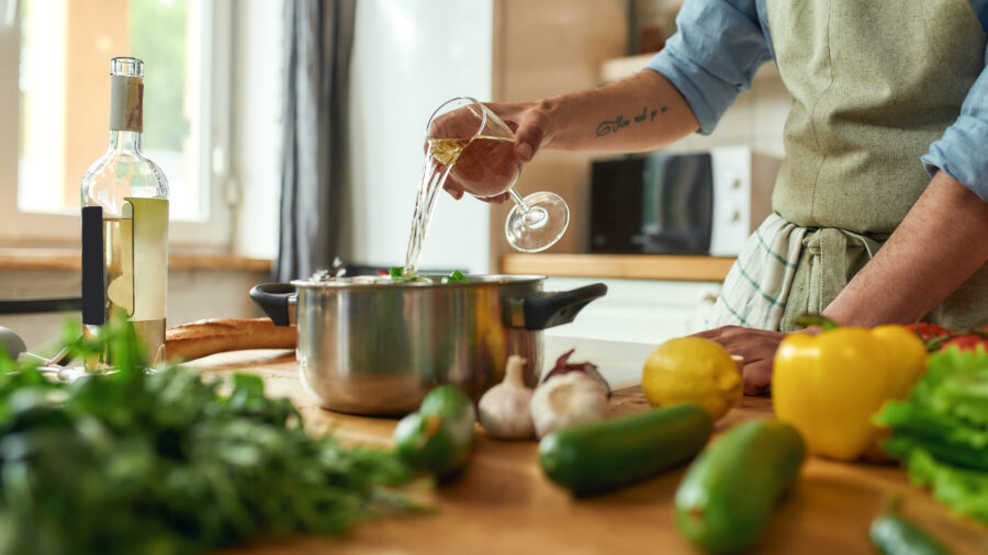 Cropped shot of man, chef cook pouring a glass of white wine into the pan with chopped vegetables while preparing a meal in the kitchen. 