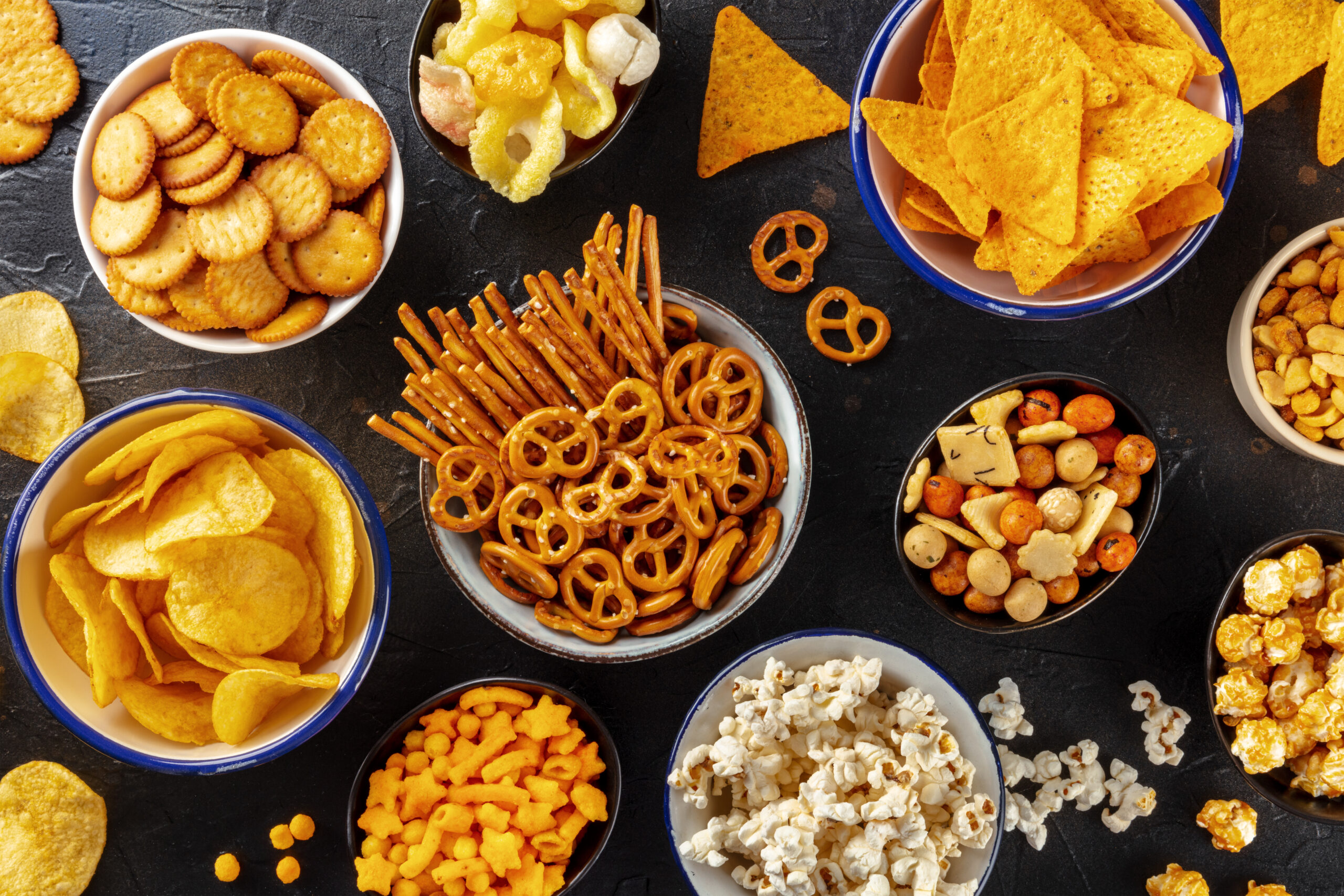 Salty snacks, party mix. An assortment of crispy appetizers, overhead flat lay shot on a black background. Potato and tortilla chips, crackers, popcorn etc