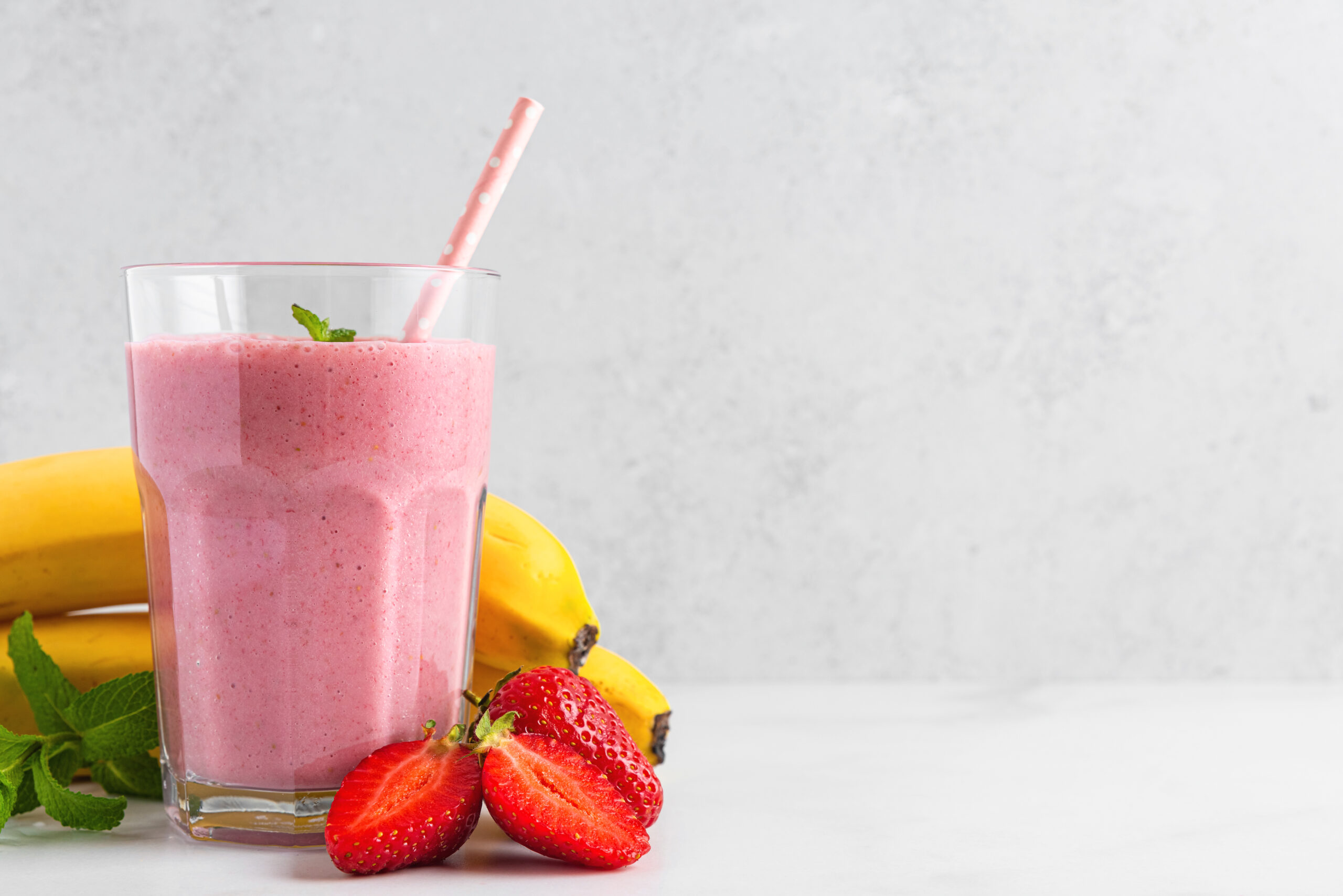 Strawberry and banana smoothie or milkshake in a glass with straw, fresh berries and mint on white background. Summer refreshing drink