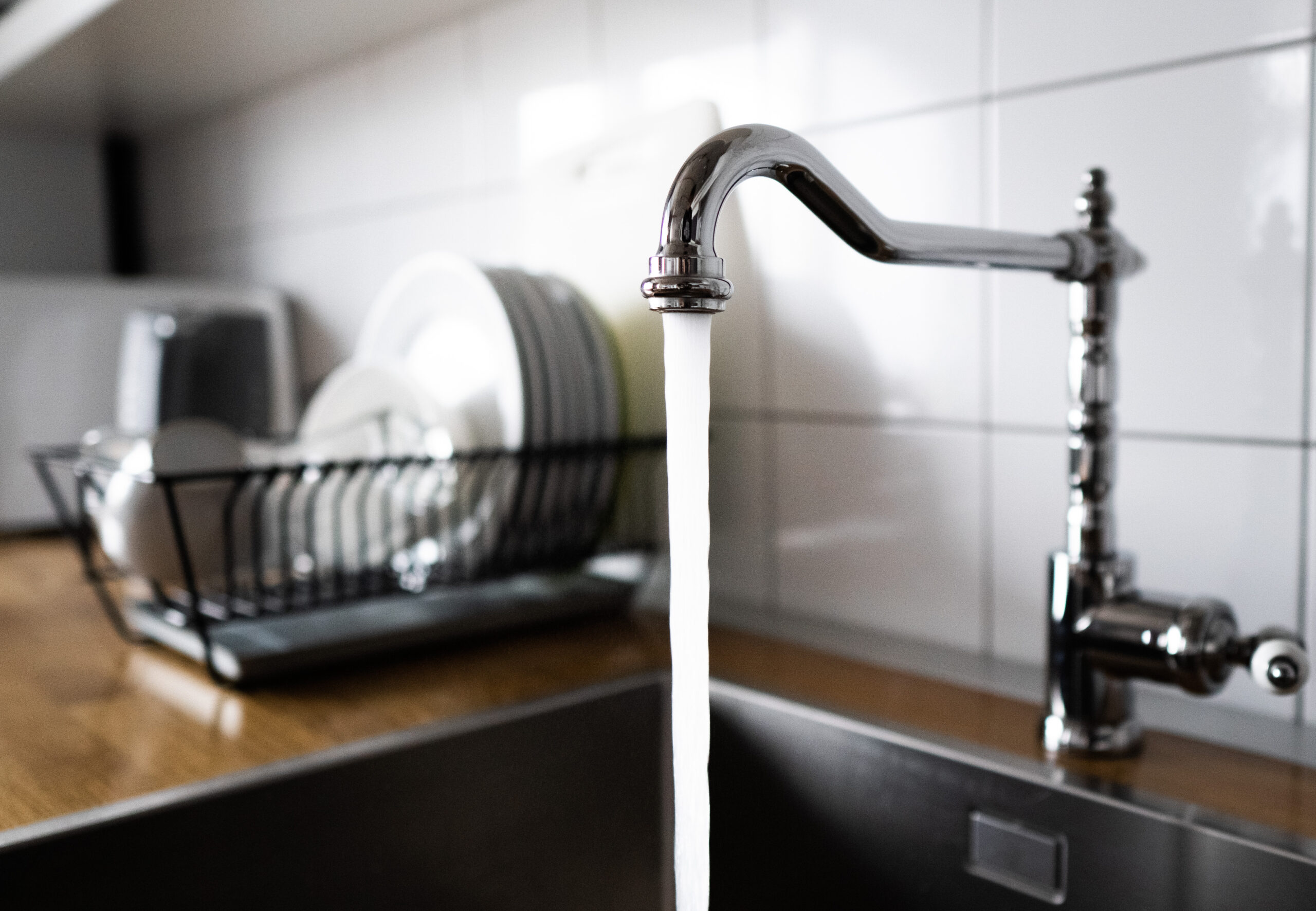 Water flowing out of a kitchen stainless steel tap into the sink. Wasting water by leaving a chrome faucet tap running. Overusing household water. Water misuse in domestic duties and activities.