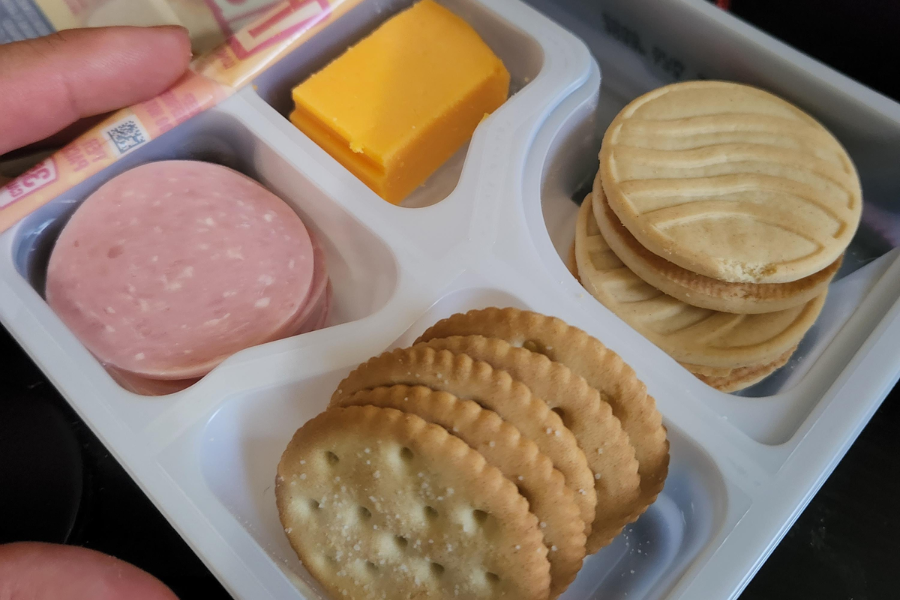 A divided plastic lunch tray containing a stack of round crackers, a few slices of deli-style meat (likely bologna), a thick slice of cheddar cheese, and two sandwich cookies with a swirl design on top.