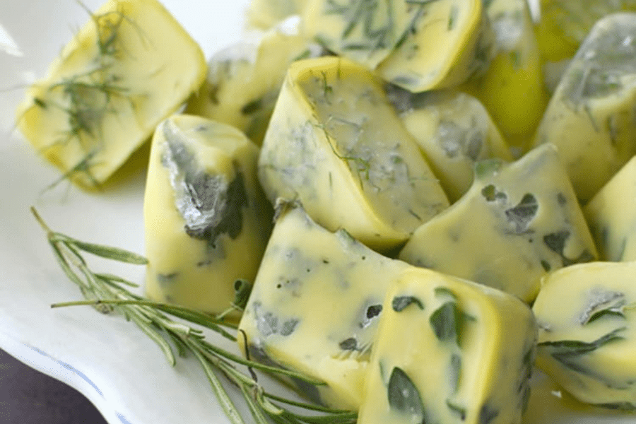 Close-up of homemade frozen herb cubes made by preserving fresh herbs in olive oil, molded in ice cube trays. The cubes show visible sprigs of rosemary, parsley, and dill suspended in golden-yellow oil, ready for easy use in cooking.