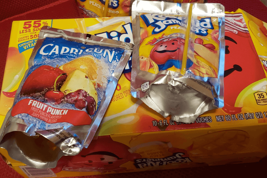 Two juice drink pouches—one Capri Sun Fruit Punch and one Kool-Aid Jammers Peach Mango—sit on top of a box of Kool-Aid Jammers. Both pouches are opened and partially flattened, showing vibrant packaging with fruit images and cartoon branding.
