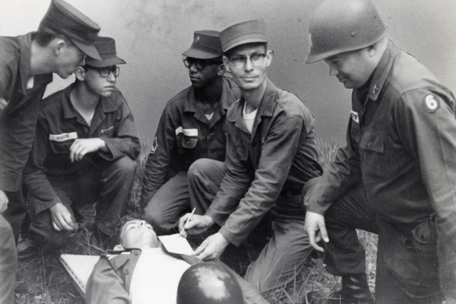 Black-and-white photo of a group of U.S. Army soldiers gathered around a medical training exercise. One soldier lies on a stretcher while others observe, as a man in glasses kneels beside the patient and writes notes on a clipboard. The team appears focused, indicating a moment of instruction or evaluation.