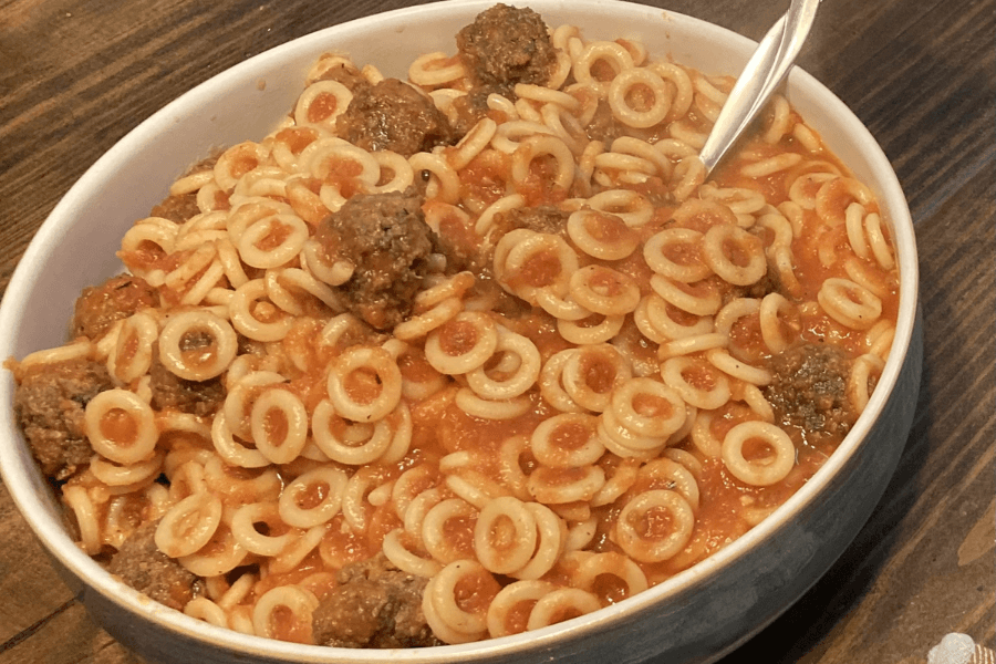 A bowl of SpaghettiOs mixed with meatballs, served in a white bowl with a spoon. The pasta rings are coated in a bright red tomato sauce and evenly mixed with small beef meatballs.