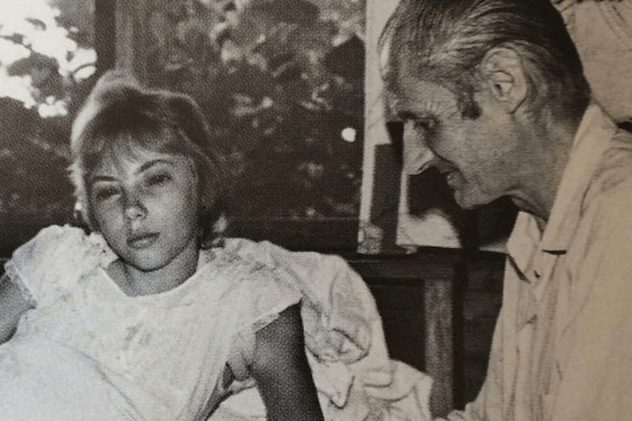 Black-and-white photo of a young woman lying in bed, appearing tired or recovering, while an older man with slicked-back hair sits beside her, smiling gently. Light filters through a window behind them, revealing blurred outdoor foliage.