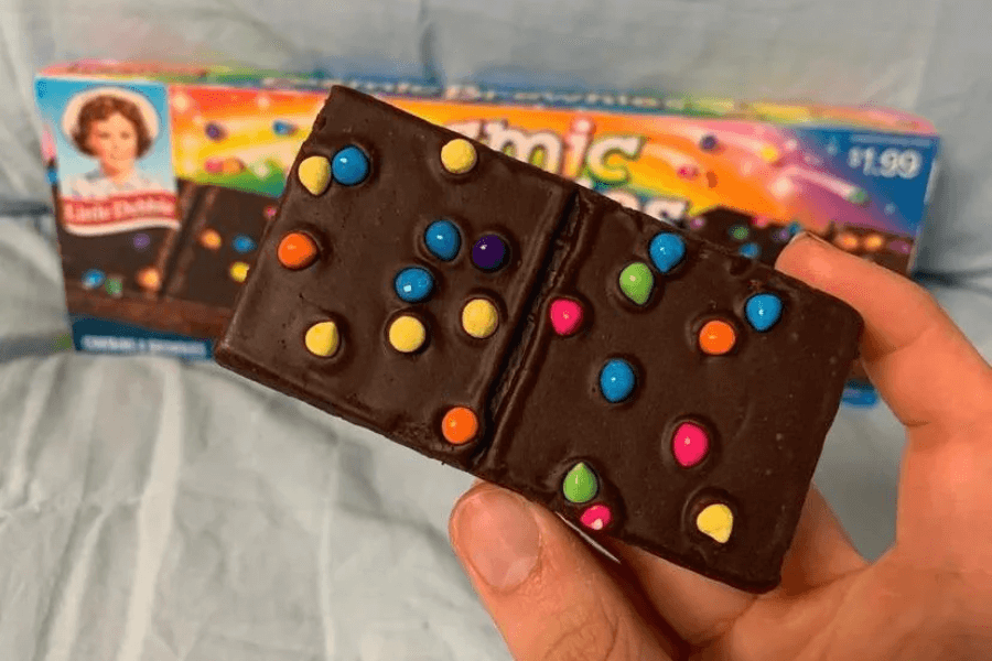 A hand holding a Little Debbie Cosmic Brownie, topped with colorful candy-coated chocolate pieces. In the background is the product’s rainbow-themed box featuring the Little Debbie logo and images of the brownies.