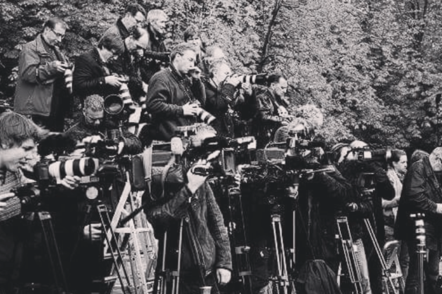 Black-and-white photo showing a large crowd of photographers and videographers, all equipped with professional cameras and long lenses, closely packed together on risers and ladders, capturing an unseen event in a wooded outdoor setting.