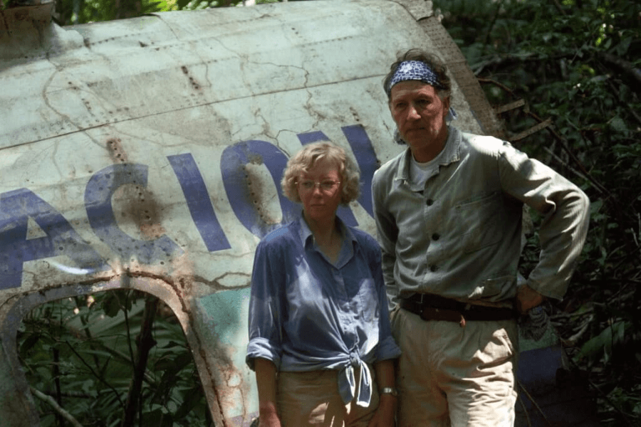 A woman and a man stand in front of a moss-covered, rusting airplane fuselage partially buried in jungle vegetation. The woman wears glasses and a light blue shirt, while the man wears a bandana and light clothing, both looking solemn and serious.