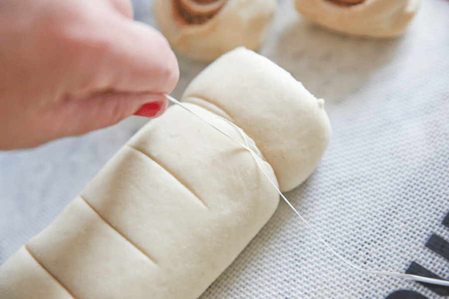 Close-up of a hand using dental floss to slice through cinnamon roll dough on a silicone baking mat. The dough has been marked for even cuts, with several rolls already sliced in the background.