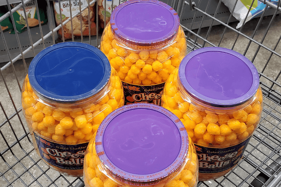 A shopping cart filled with five large plastic containers of Utz baked cheddar cheese balls. Four containers have purple lids and one has a blue lid, all filled with bright orange cheese-flavored snacks.
