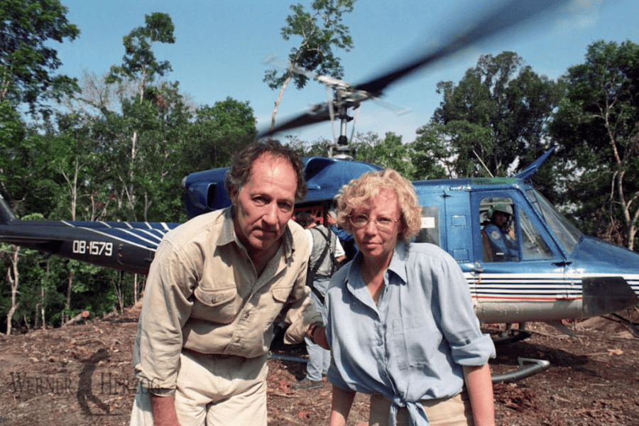 A man and woman in outdoor expedition clothing stand in front of a blue helicopter with white and red stripes, landed in a jungle clearing. The woman wears glasses and a light blue shirt, while the man leans slightly forward toward the camera. The helicopter’s rotor blades are spinning, and a pilot is visible inside.