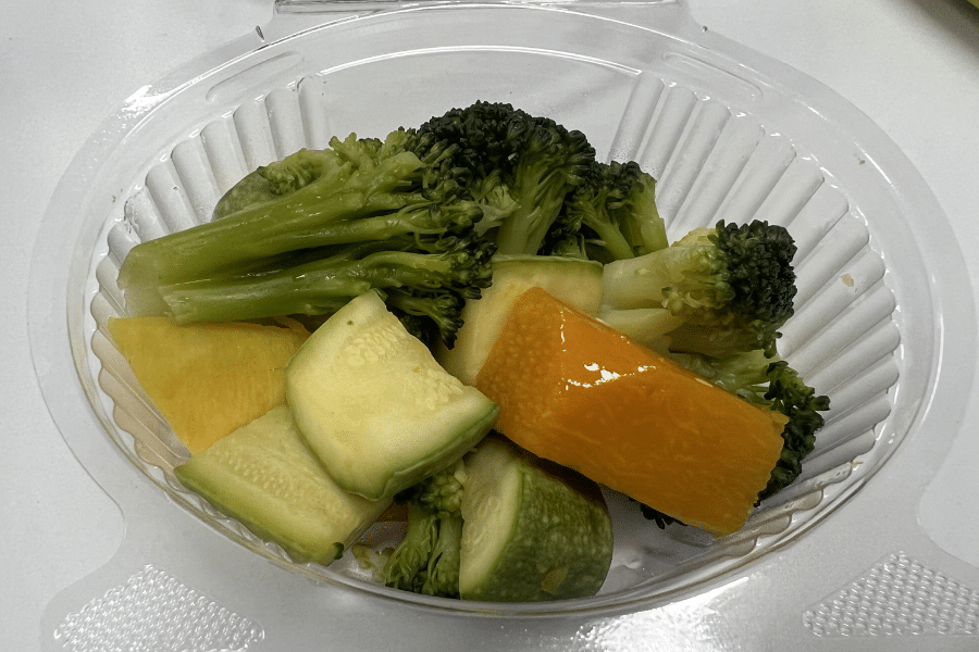 Plastic takeout container filled with a medley of steamed vegetables including broccoli florets, yellow bell pepper, zucchini slices, and chunks of yellow squash, arranged on a white surface.