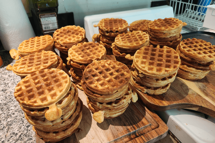 Multiple tall stacks of golden brown waffles are arranged on wooden cutting boards and a kitchen counter. The waffles have a crisp, grid-patterned surface and are stacked in varying heights, suggesting a large batch was freshly made.