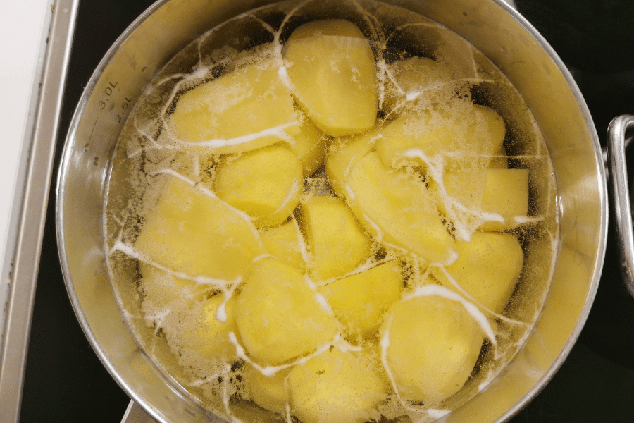 Top-down view of a stainless steel pot filled with peeled potatoes boiling in water, with white foam forming on the surface from starch release. The pot is on a stovetop and marked with volume measurements inside.