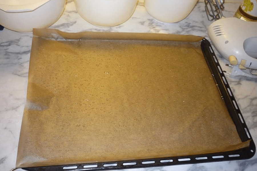 Baking sheet lined with parchment paper that has been lightly sprinkled with water droplets, likely to prevent sticking or help shape the paper to the tray. The tray is on a marble countertop surrounded by kitchen tools and mixing bowls.