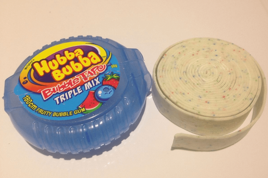 A blue plastic container of Hubba Bubba Bubble Tape Triple Mix bubble gum sits next to the rolled-out gum, which is white with multicolored candy-like specks. The packaging features fruit graphics and bold, colorful branding.