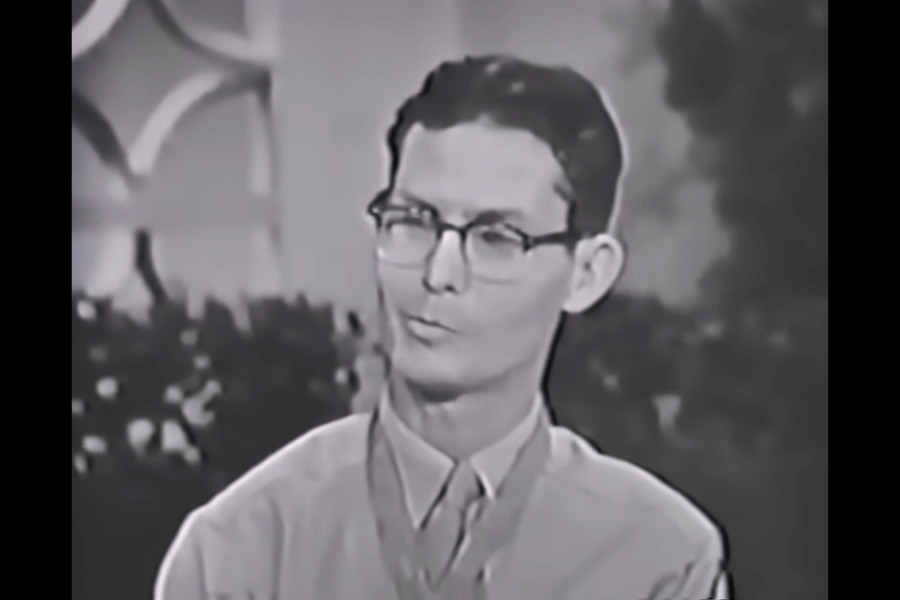 Black-and-white still from a television interview showing Desmond Doss wearing glasses and a collared shirt, with the Medal of Honor around his neck. He appears calm and composed while speaking, with a softly lit background featuring plants and abstract patterns.