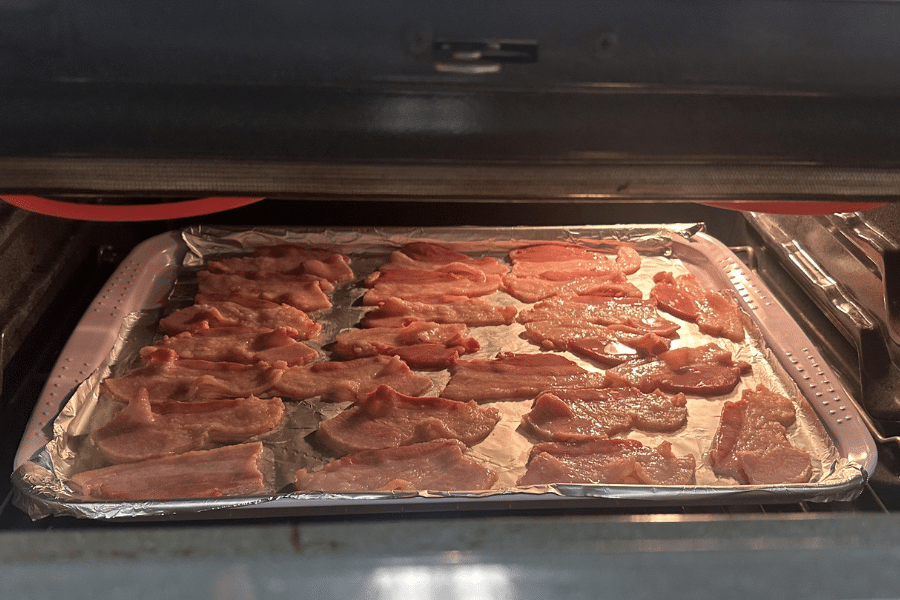View inside an oven showing multiple strips of raw bacon laid out on a foil-lined baking sheet, arranged in rows for even cooking. The bacon is beginning to sizzle as the oven heats.