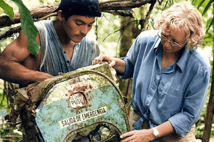 A man and a woman inspect a rusted and moss-covered airplane emergency exit door labeled “SALIDA DE EMERGENCIA” in the middle of a dense jungle. The woman wears glasses and a blue shirt, and the man, in a sleeveless top, examines the wreckage closely.