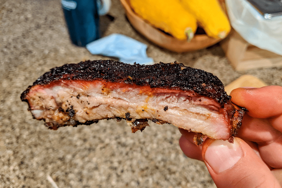 Close-up of a hand holding a smoked pork rib with a dark bark crust, visible pink smoke ring, and juicy interior layers of fat and meat. The background shows a kitchen counter with yellow squash, a blue cup, and a blue face mask.