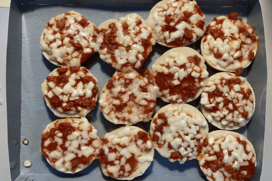 A baking tray filled with frozen mini pizzas, each topped with small diced pepperoni pieces and melted mozzarella cheese over tomato sauce, arranged in a slightly overlapping pattern.