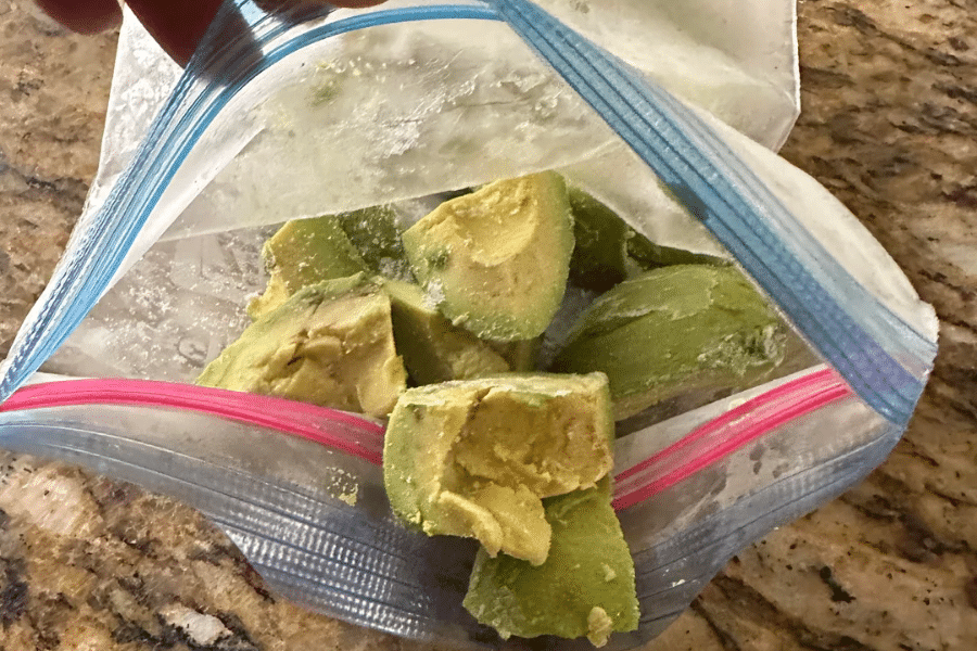 Chunks of frozen avocado stored in a resealable freezer bag, shown on a kitchen countertop. The avocado pieces are slightly frosty and appear ready for smoothies or future use in recipes.