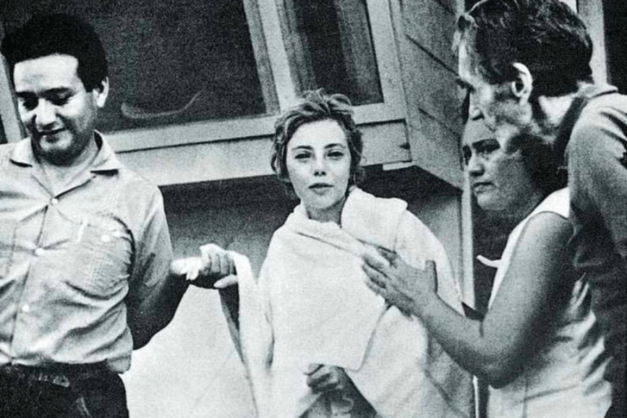 Black-and-white photo of a young woman wrapped in a towel being supported by three concerned adults, two men and one woman. She appears calm but visibly exhausted, likely in a moment of recovery or rescue, standing outside a building.