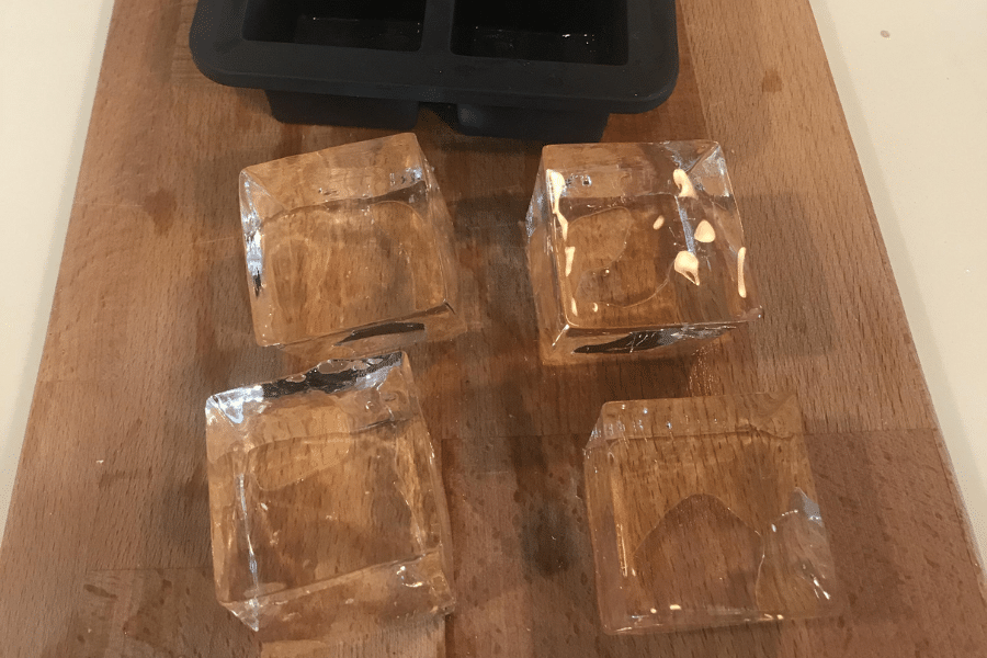 Four perfectly clear square ice cubes placed on a wooden cutting board, with a black silicone mold tray in the background. The ice cubes are crystal clear, indicating slow or directional freezing methods.