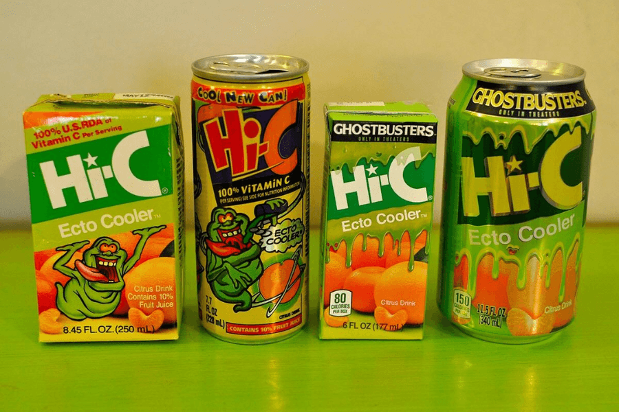 Four different Hi-C Ecto Cooler citrus drink packages, including two juice boxes and two cans, are lined up on a green surface. Each features bright green and orange designs, with some versions showing the Ghostbusters logo and the Slimer character from the franchise.