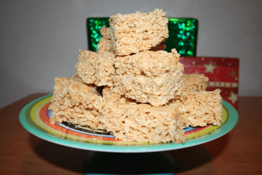 A colorful cake stand stacked with homemade Rice Krispies treats cut into uniform rectangular bars. The background includes shiny green and red holiday-themed gift boxes, adding a festive touch.