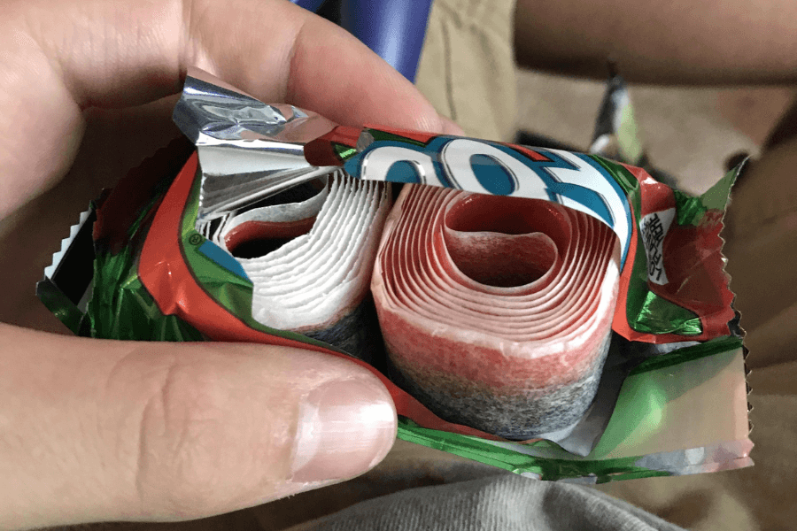 A close-up of an unwrapped Fruit by the Foot candy roll held in someone’s hand. The rolled strip of red, white, and black fruit-flavored candy is partially unraveled, revealing its layered, flexible texture inside the shiny foil wrapper.