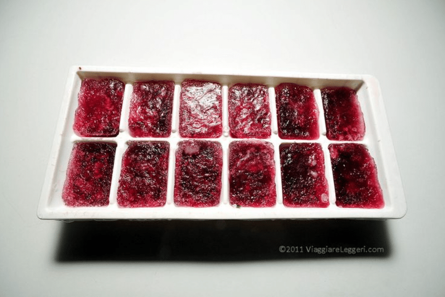 White plastic ice cube tray filled with frozen dark red cubes, likely made from blended or juiced berries. The tray is set on a plain white surface under direct lighting, highlighting the textured surface of each cube.