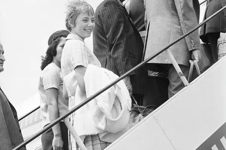 Black-and-white photo of a smiling young woman boarding an airplane via a stairway, surrounded by other passengers in formal clothing. She carries a folded blanket or coat, looking back toward the camera as she ascends.