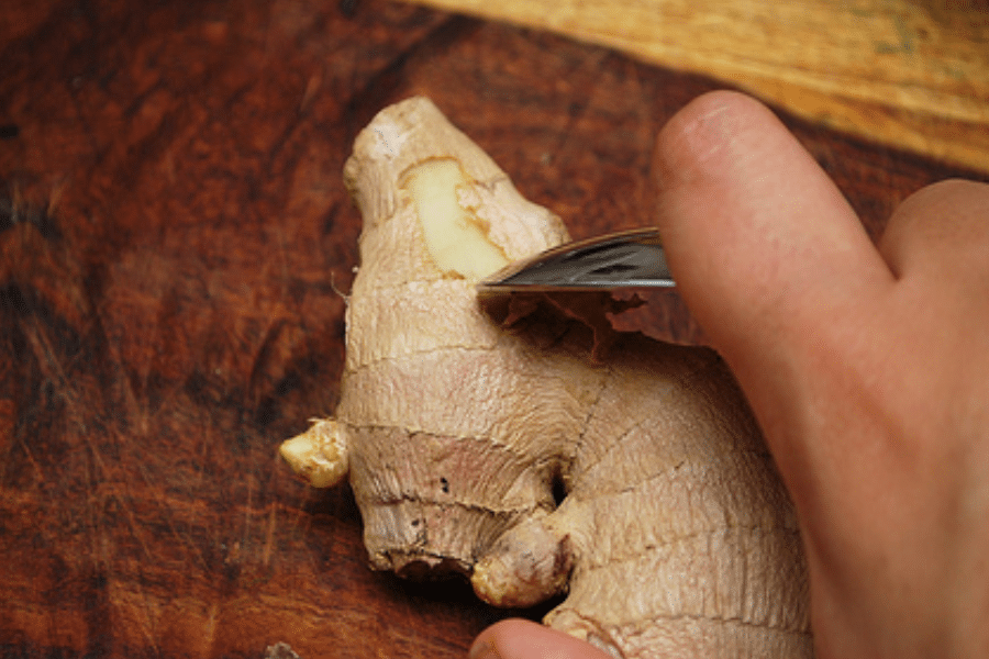 Close-up of a hand using a spoon to peel the skin off a fresh ginger root on a dark wooden cutting board. The spoon gently scrapes the thin skin away, revealing the pale yellow flesh underneath.