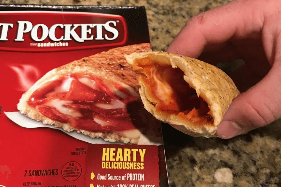 A hand holds a bitten-into Hot Pocket stuffed with melted cheese and pepperoni in tomato sauce, revealing its gooey filling. In the background is the red Hot Pockets box showing an image of the same sandwich and product information like "Hearty Deliciousness" and "Good Source of Protein."
