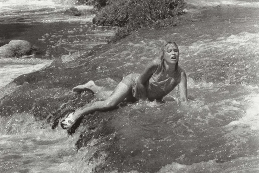 Black-and-white photo of a young woman struggling to move through a fast-flowing stream or river, visibly distressed as she braces herself against the current with one hand. Her clothing is soaked, and her body language conveys exhaustion and urgency.