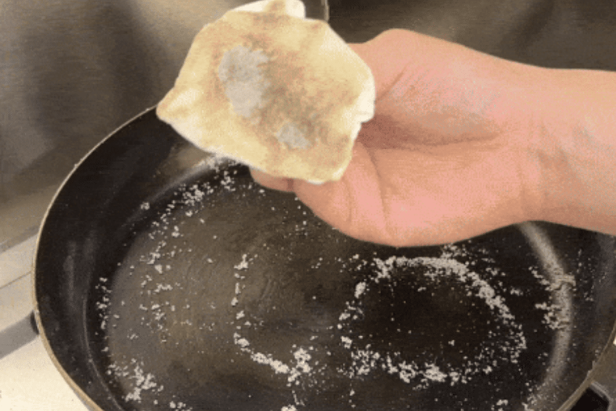 A hand holding a folded paper towel showing grease and grime lifted from a frying pan, which has coarse salt sprinkled inside. The salt is being used as a natural abrasive to scrub the pan.
