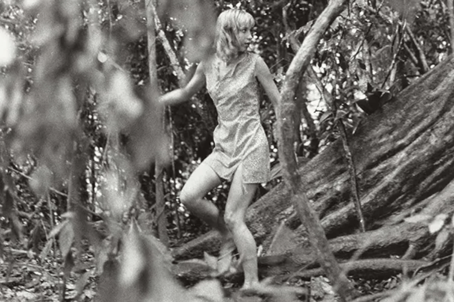 Black-and-white photo of a young woman cautiously moving through a dense jungle, barefoot and wearing a sleeveless dress. She grips vines for support while stepping over roots and uneven terrain, glancing behind her with a tense expression.