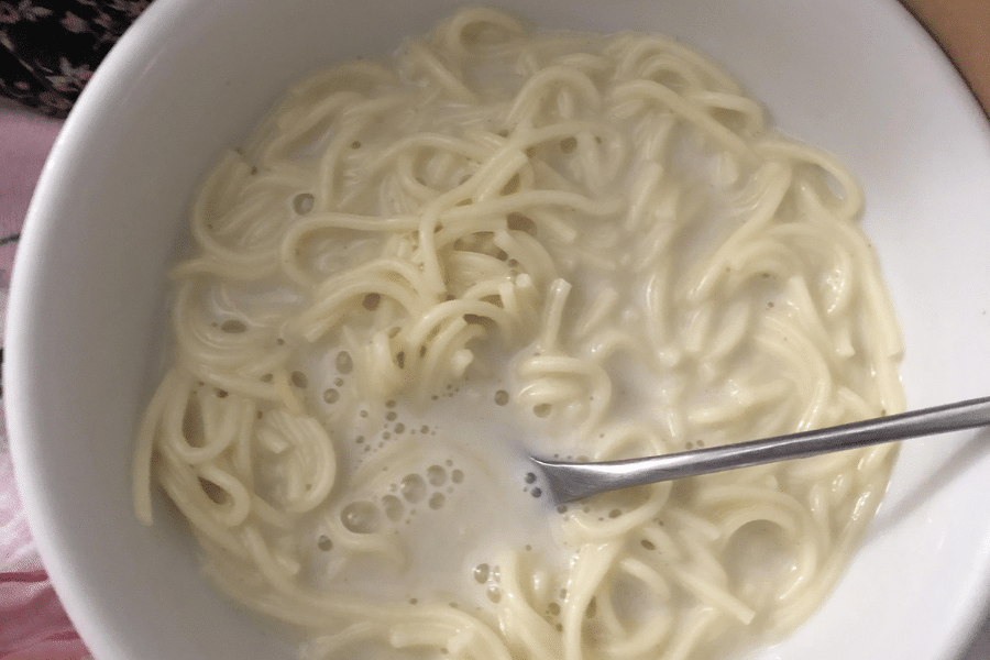 Bowl of hot milk noodles, featuring soft cooked spaghetti submerged in a creamy milk broth with a spoon resting inside. The dish appears simple and comforting, often served as a warm snack or breakfast.