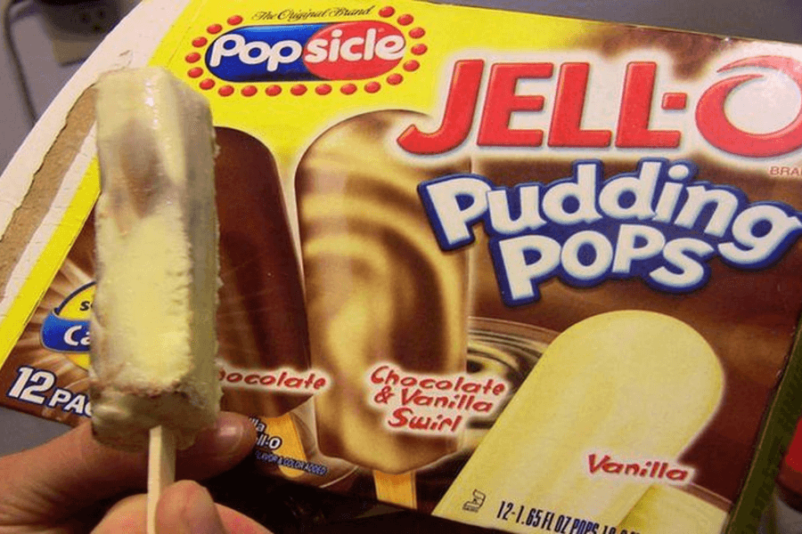A hand holds a partially eaten chocolate and vanilla swirl JELL-O Pudding Pop in front of the product’s packaging. The box shows three flavors—chocolate, chocolate & vanilla swirl, and vanilla—and is branded with the Popsicle and JELL-O logos.