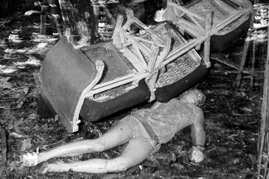 Black-and-white reenactment photo of a woman lying unconscious on the jungle floor beneath overturned airplane seats. Her dress is torn and her body appears bruised, depicting the immediate aftermath of a plane crash.