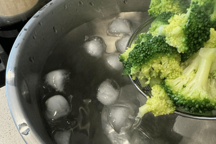 Freshly blanched broccoli being lowered into a large bowl of ice water using a strainer, with visible ice cubes floating to stop the cooking process. The ice bath technique helps preserve the broccoli’s bright green color and crisp texture.