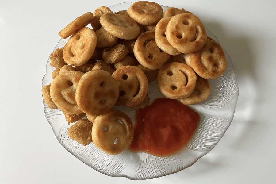 A clear glass plate filled with golden-brown smiley face fries and small breaded nuggets, served with a generous portion of ketchup on the side. The fries have a crispy exterior and are shaped with cut-out eyes and smiles.