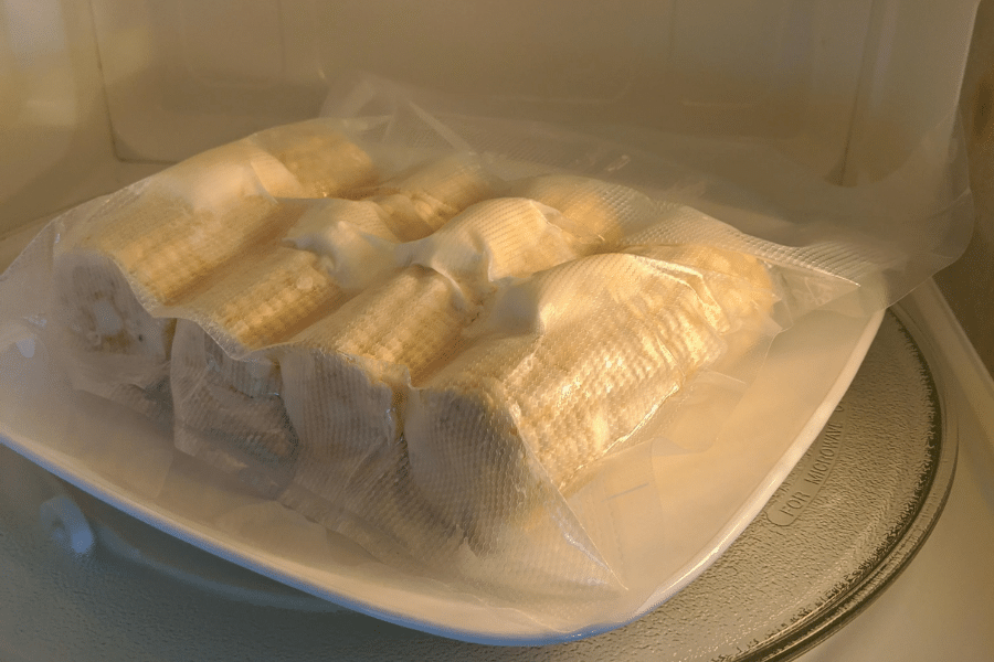 Vacuum-sealed bag of corn on the cob placed on a white plate inside a microwave, prepared for steaming or reheating. The corn is tightly packed and appears partially cooked.