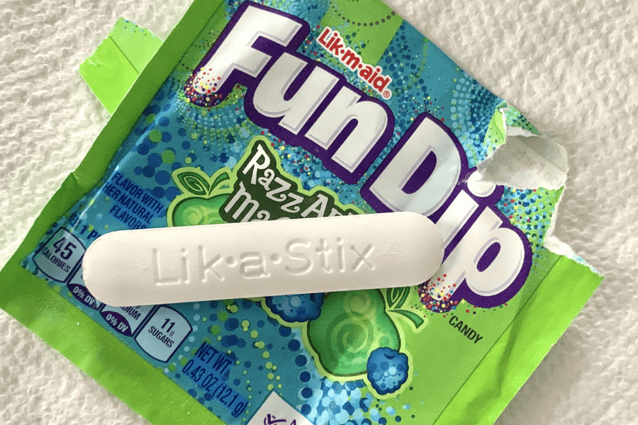 A torn Fun Dip candy packet in the RazzApple Magic flavor is shown with a white candy stick labeled “Lik-a-Stix” resting on top. The packaging is bright green and blue with berry and apple graphics, sitting on a textured white surface.